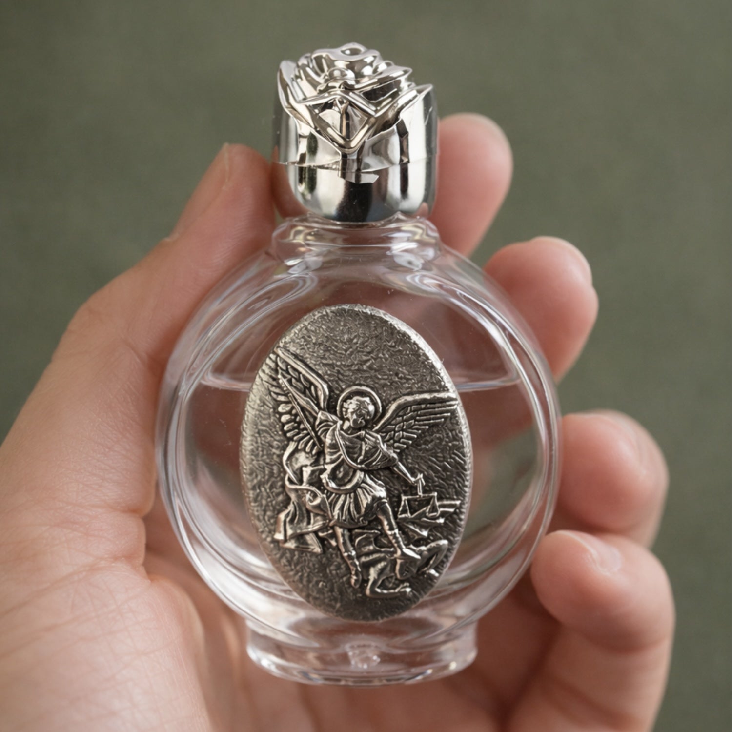 Clear glass bottle with silver angel emblem held in a hand against a neutral background